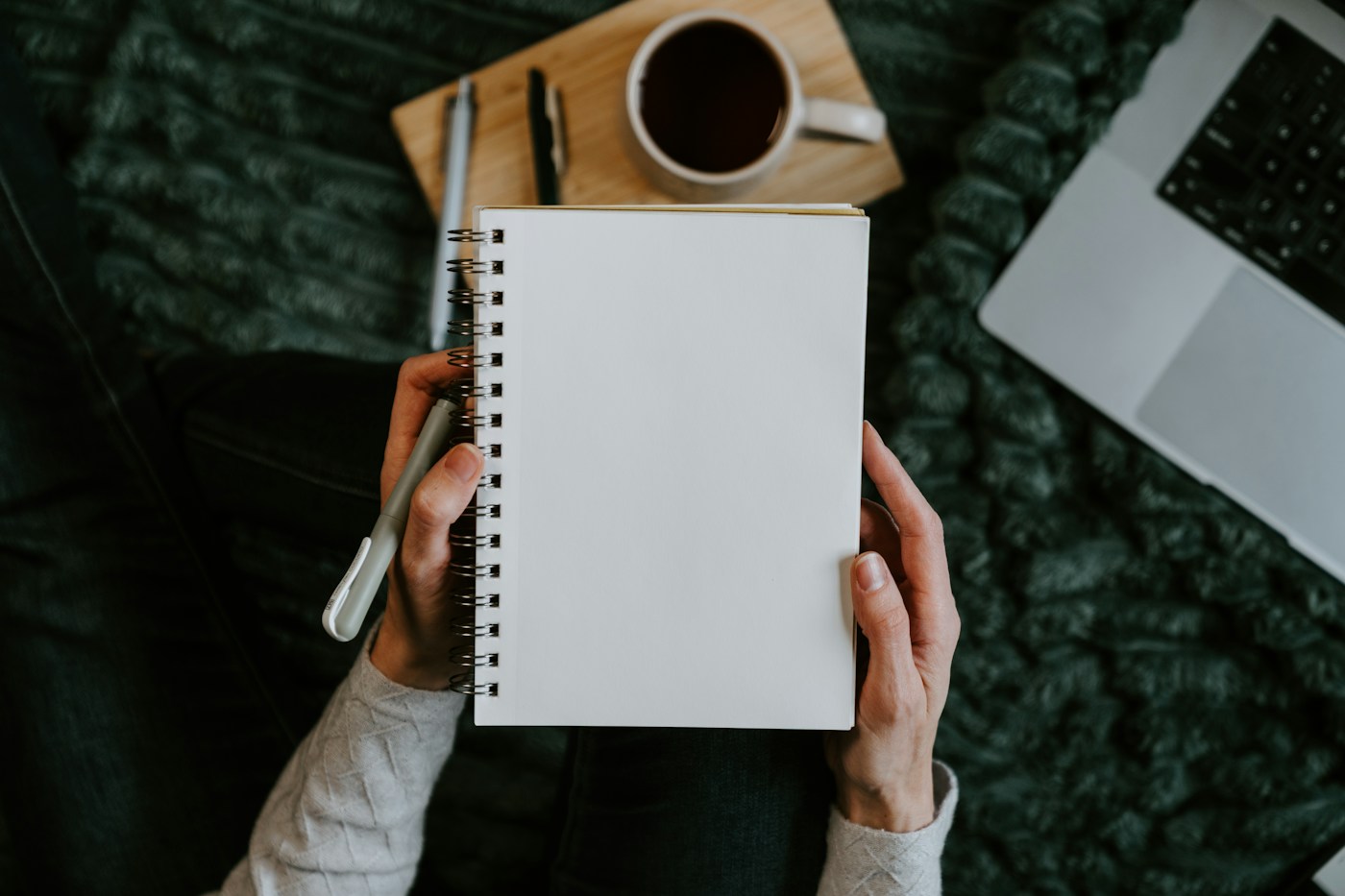 Manos sosteniendo una libreta en blanco junto a un portátil y una taza de café