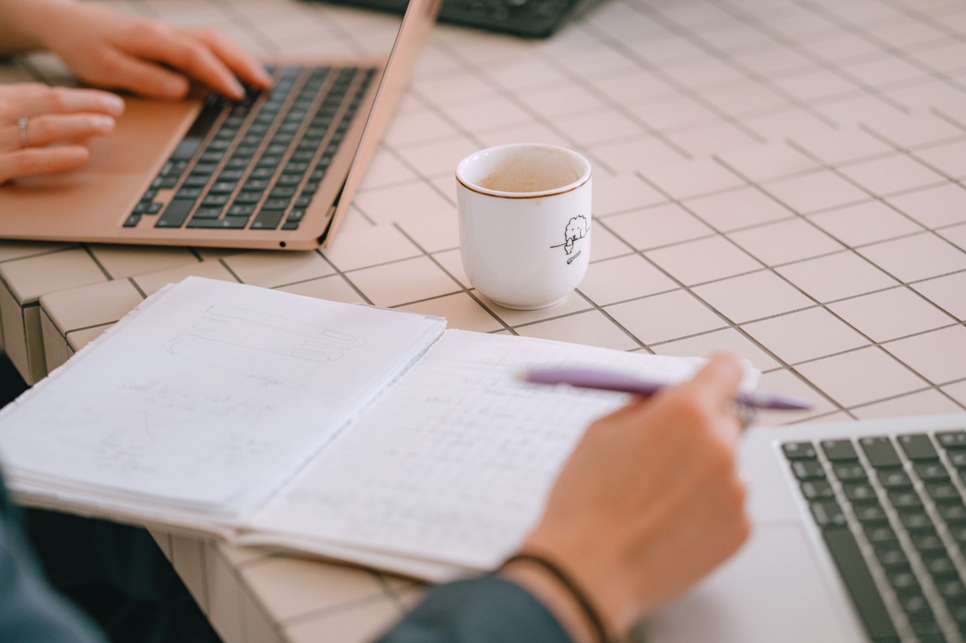 Dos personas trabajando con portátiles y una libreta sobre una mesa de azulejos claros