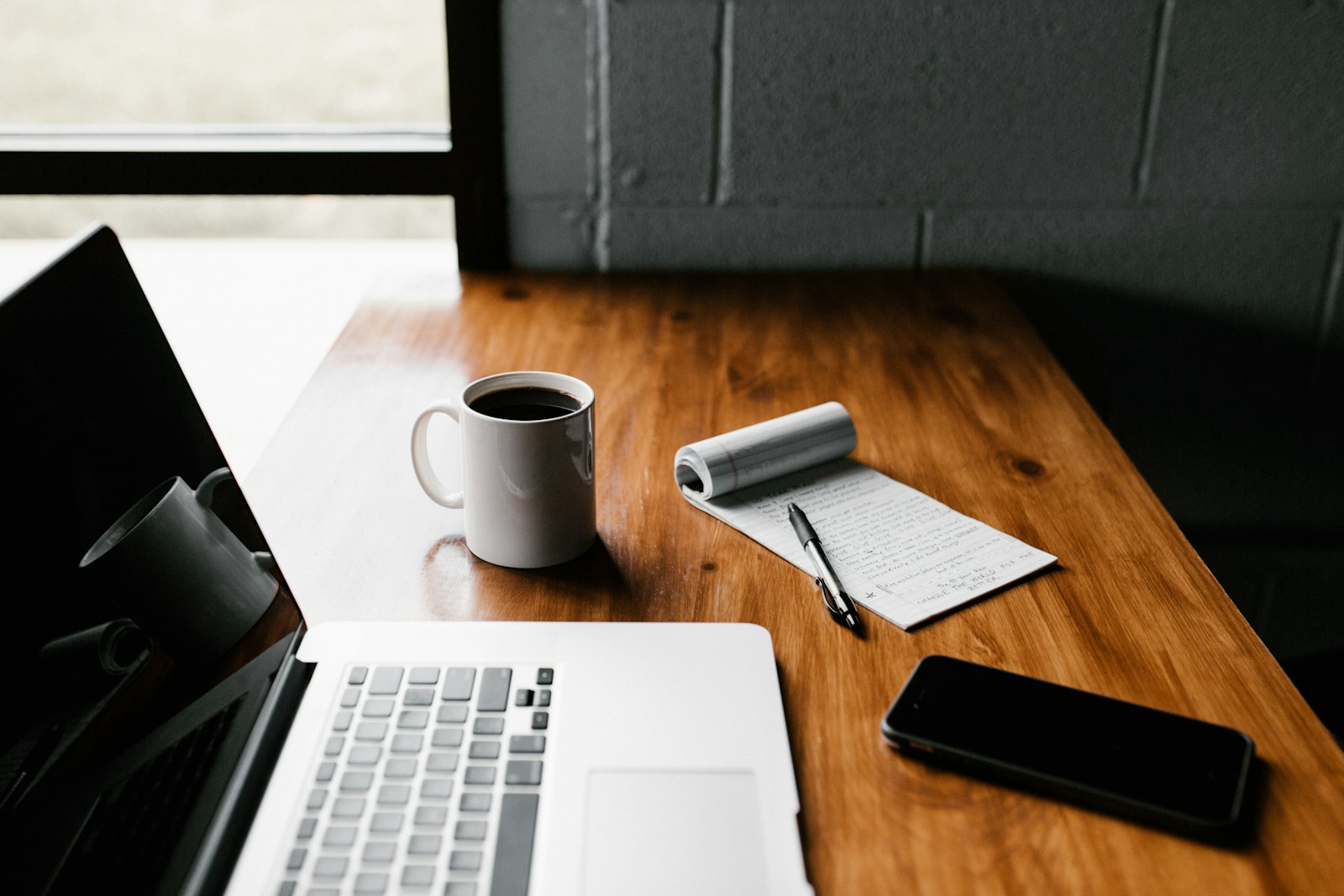 Computadora portátil, teléfono, libreta y taza de café sobre una mesa de madera.